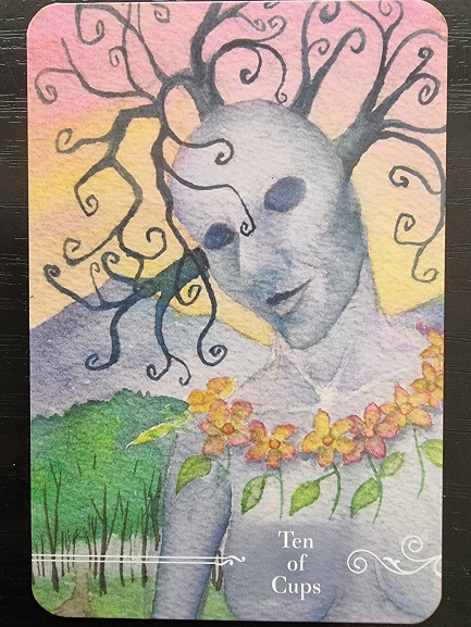 A surreal tarot card from the Guardian Tarot, the Ten of Cups features a blue-gray, humanoid figure with tree-branch hair and hollow eyes, adorned with a garland of orange flowers against a colorful sky and green landscape.