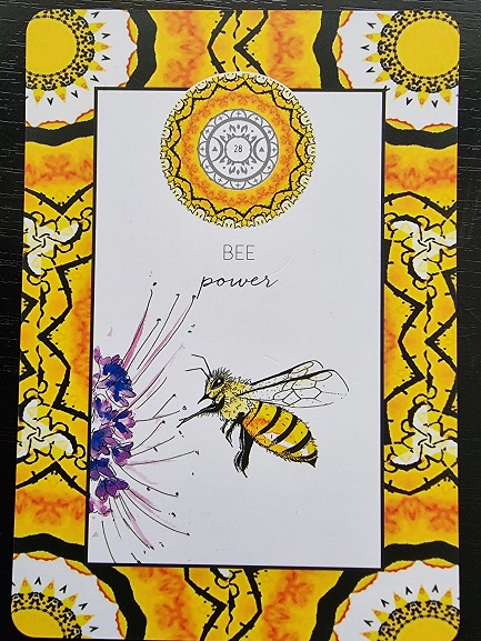 A tarot-style card from the Animal Kin Oracle features a bee approaching a purple flower, with the words BEE and power above. The border displays a yellow and black geometric pattern with circular sun-like designs. Card number is 28.