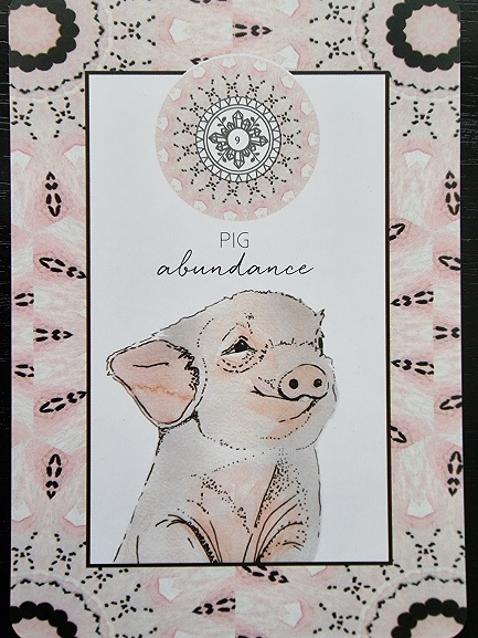 A card from the Animal Kin Oracle featuring an illustrated pig with the word “abundance” above it. The background showcases a pink and black symmetrical pattern, a decorative circular design, and the text: “Pig abundance.”.