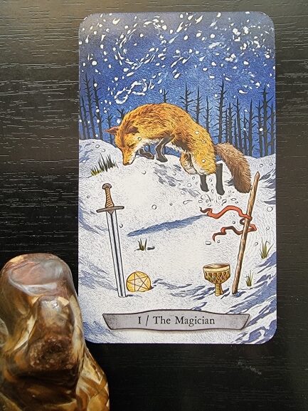 A tarot card titled The Magician shows a fox in a snowy landscape. In front of it lie a sword, a wand with a red ribbon, a pentacle, and a cup. A carved brown stone figure sits nearby on a dark surface.