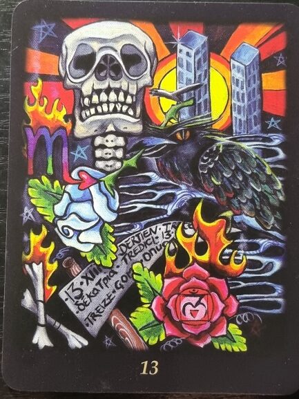 A colorful Bonefire Tarot-style card features a skull, twin towers, a black crow, flames, a white and a red rose, a purple Scorpio symbol, and a yellow sun. There is also a note with writing and the number 13 at the bottom.