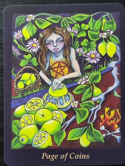A tarot card illustration from the Bonefire Tarot, titled Page of Coins, shows a girl with long hair and purple eyes squeezing lemons. She’s surrounded by vibrant green leaves, flowers, sliced lemons, a fire, and a basket of coins.