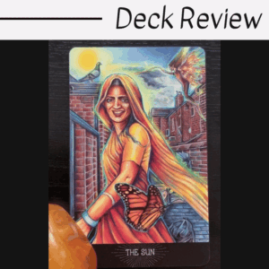 A tarot card from the Everyday Enchantment Tarot titled The Sun shows a smiling woman in an orange sari with a butterfly on her hand, birds above, and brick buildings behind her. An orange crystal rests on the card’s corner. Deck Review is written above.