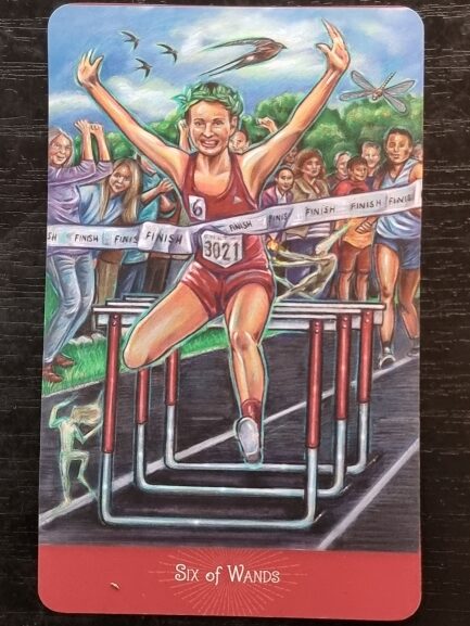 A triumphant runner leaps over the final hurdle, arms raised in victory, breaking the finish line tape. Cheering spectators and competitors are in the background, celebrating this tarot-inspired moment—perfect for reflection or journaling. The card is titled Six of Wands.
