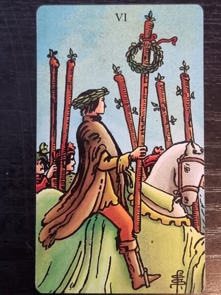 A tarot card shows a person riding a white horse, holding a wand adorned with a wreath. Others hold wands in the background, suggesting a parade or celebration—perfect imagery for insightful tarot and journaling sessions. The card is labeled VI at the top.