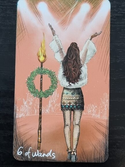 A tarot card illustration of a woman standing on stage with arms raised, perfect for tarot and journaling. She faces away in a short skirt and fluffy top, beside a decorated wand with a wreath under spotlights. The card reads 6 of Wands.