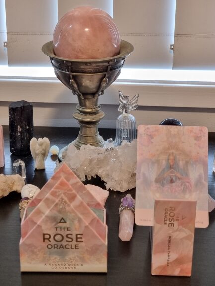 A display of crystals, including rose quartz, angel figurines, and The Rose Oracle card deck, is arranged on a dark surface in front of a window with blinds. A large pink sphere sits in an ornate silver stand beside The Rose Oracle.