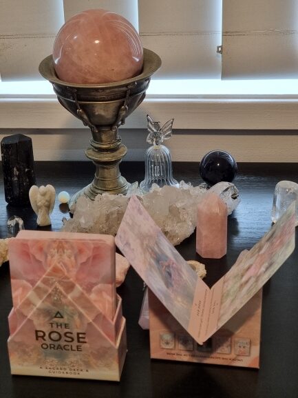 A display of crystals, featuring a large rose quartz sphere on a stand, smaller crystals, angel figurines, and The Rose Oracle cards opened and standing gracefully on a dark surface.