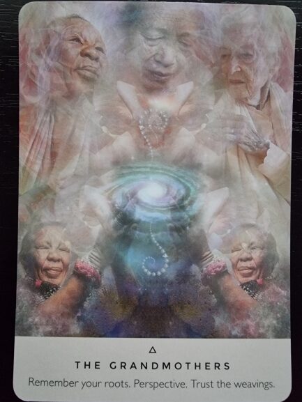 A tarot card titled The Grandmothers from The Rose Oracle shows ethereal images of elderly women surrounding a glowing, swirling galaxy held in cupped hands. Text reads: Remember your roots. Perspective. Trust the weavings.