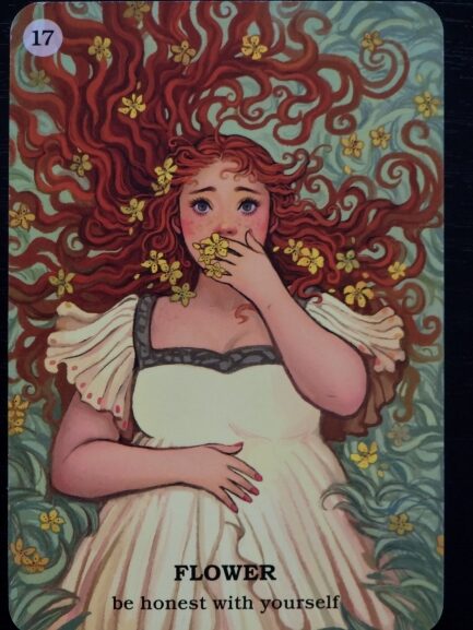 An illustrated card from Believe In Your Own Magic shows a woman with long, flowing red hair adorned with yellow flowers. She wears a white dress and holds flowers to her mouth. Text below reads: FLOWER – be honest with yourself.