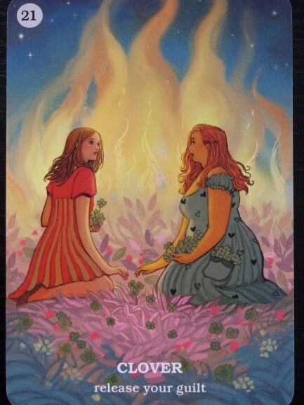 Two women kneel on pink grass, holding clovers and facing each other before a glowing fire. The card reads, CLOVER release your guilt, number 21 in the corner—a reminder to Believe In Your Own Magic.