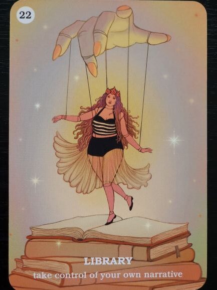 Illustration of a woman as a marionette puppet controlled by a large hand above, standing on an open book with stacked books below. Text reads: LIBRARY—Believe In Your Own Magic and take control of your own narrative.