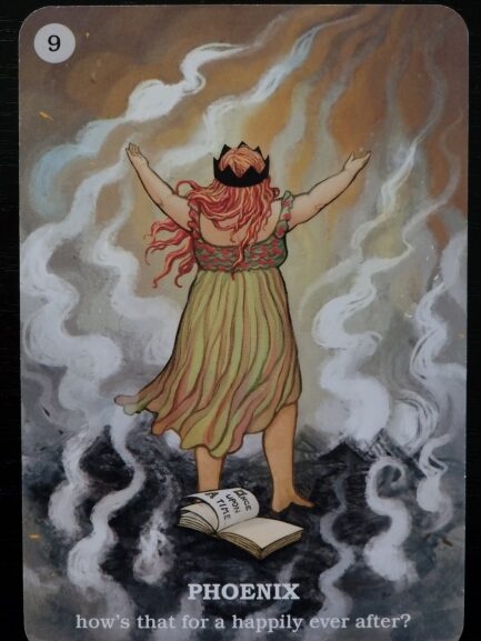 A woman with red hair and a crown stands with arms raised amid swirling smoke and flames. An open book lies at her feet. Text reads: PHOENIX – Believe In Your Own Magic. How’s that for a happily ever after? Number 9 is in the top left corner.