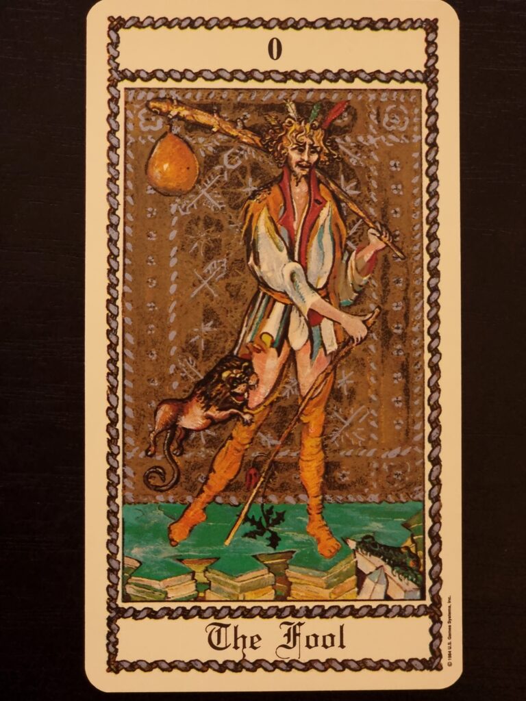 A tarot card from The Medieval Scapini Tarot titled Two of Coins features a seated figure with two coins, surrounded by ornate red drapery, symbols, and two robed figures above, all set against a cream background.