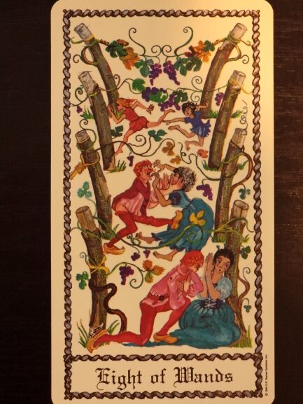 A colorful tarot card from The Medieval Scapini Tarot titled Eight of Wands shows eight wooden wands surrounded by green vines and purple grapes, with several people interacting playfully among the wands and flowers.
