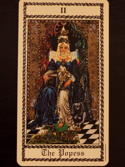 A tarot card from The Medieval Scapini Tarot titled The Popess shows a crowned woman in blue and white robes, seated on a throne with a book in her lap, flanked by black and white pillars, and a crescent moon at her feet.