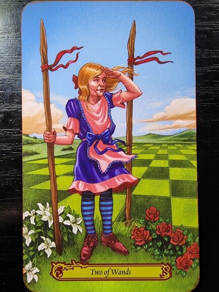 A young girl in a pink and blue dress stands between two wands on a checkered green field in Tarot in Wonderland, surrounded by white lilies and red roses. She gazes into the distance with one hand shielding her eyes. The card reads Two of Wands.