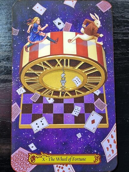 An illustrated Tarot in Wonderland card shows Alice and the White Rabbit running on a large, floating clock face. Playing cards and a chessboard swirl around them against a purple, starry background. Label reads “X – The Wheel of Fortune.”.