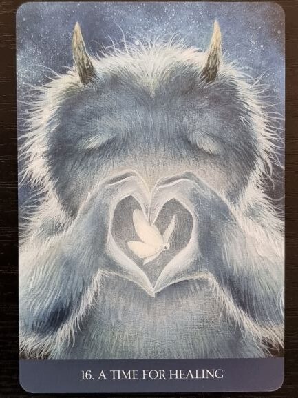 A gentle, furry creature with small horns closes its eyes and forms a heart shape with its hands, framing a glowing white butterfly inside. From Into the Lonely Woods, the card reads, 16. A TIME FOR HEALING.