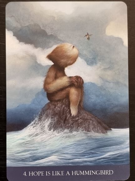 A furry creature sits on a rock surrounded by water, gazing up at a small hummingbird flying in the cloudy sky above. Into the Lonely Woods, hope is like a hummingbird.