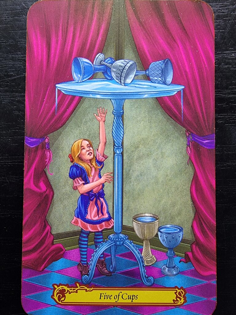 A young girl in a blue and pink dress reaches for tipped-over cups on a tall table. Liquid spills from three cups, while two upright cups sit on the floor. Red curtains frame this Tarot in Wonderland scene. The card reads Five of Cups.