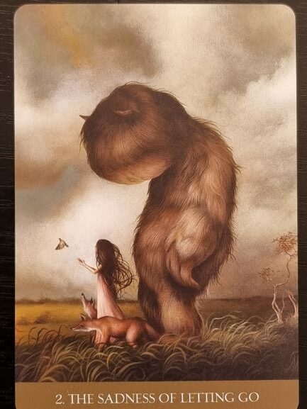 A girl stands in a field with a large, sad, furry creature beside her and a fox at her feet. She reaches toward a butterfly under a cloudy sky. Text below reads, 2. THE SADNESS OF LETTING GO – Into the Lonely Woods.