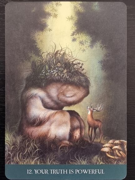 A large, earthy creature with plants growing from its head sits curled on the ground, facing a small, alert deer. Sunlight filters through trees above, evoking the quiet magic of Into the Lonely Woods. Text at the bottom reads, 12. YOUR TRUTH IS POWERFUL.