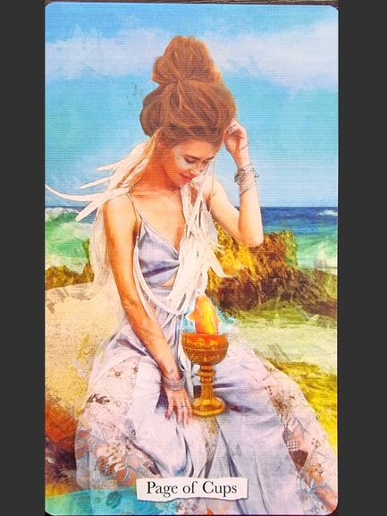 A woman in a flowing light dress sits on rocks by the ocean, holding a golden cup with a fish inside. The bright blue sky above and "Page of Cups" below complete this enchanting Superlunaris Tarot scene.