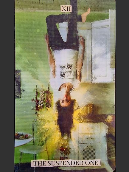 A person hangs upside down by one foot in a kitchen, glowing with yellow light. The card reads XII at the top and THE SUSPENDED ONE at the bottom, inspired by the modern Superlunaris Tarot deck’s take on the Hanged Man.
