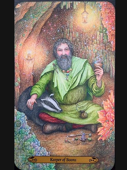 A bearded man in a green robe sits in a cozy, glowing cave, writing beside a resting badger. He holds a cup, surrounded by crystals, candles, and a lantern. The card reads Keeper of Boons.
