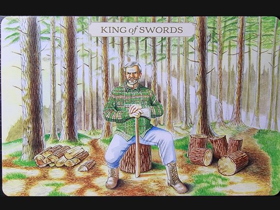 A man in a green plaid shirt and boots sits on a tree stump in a forest, holding a sword upright. Stacked and scattered logs surround him. The card reads King of Swords at the top, from the Pastoral Tarot deck.