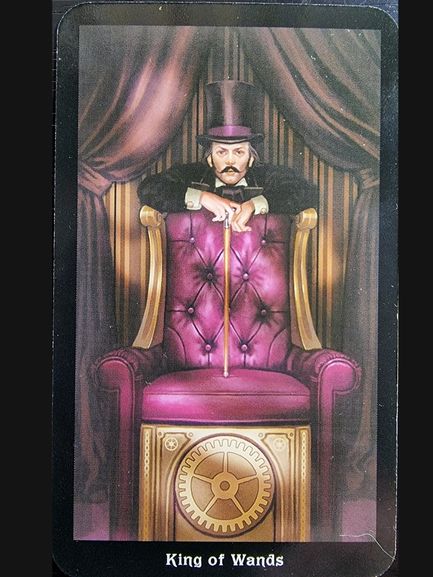 A man in a top hat and suit leans on a golden cane while sitting on a tall, purple throne adorned with gears; the card reads King of Wands at the bottom. Steampunk curtains hang in the background, inspired by The Steampunk Tarot.
