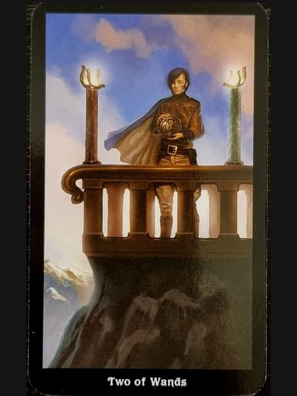 A person stands on a stone balcony holding a globe, gazing over mountains. Two upright wands frame the scene beneath a partly cloudy sky. Inspired by The Steampunk Tarot, the card reads Two of Wands.