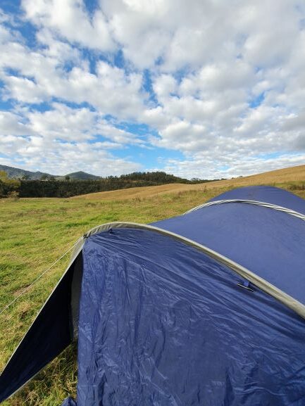 A blue tent is set up on grassy land, immersed in nature under a partly cloudy sky, with rolling hills and distant trees in the background.