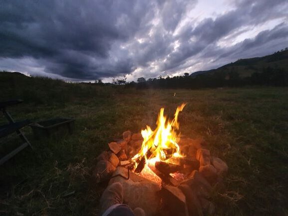 A campfire burns brightly in a stone fire ring on grassy ground at dusk, surrounded by the beauty of nature as dark clouds gather overhead and rolling hills stretch into the distance.