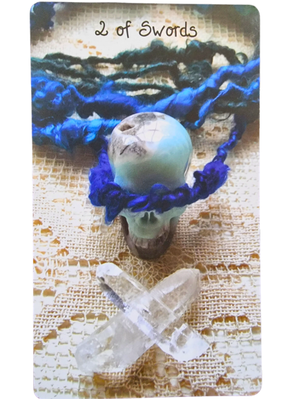 A Crystal Skull Tarot card labeled 2 of Swords shows a small skull wrapped with blue yarn and a cross of two clear crystals below, all resting on a textured, woven surface.