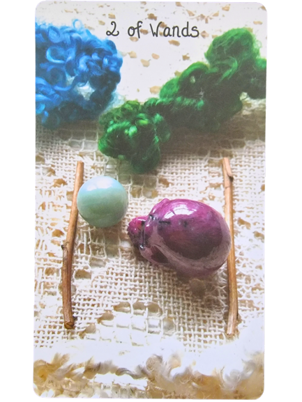 A handmade 2 of Wands card from the Crystal Skull Tarot shows two wooden sticks on lace fabric, with a green bead and a purple skull bead between them, and blue and green yarn in the background.