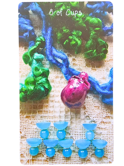 A tarot card from the Crystal Skull Tarot labeled 8 of Cups shows eight small blue cups in two rows, colorful textured yarn, and a shiny pink pouch on a woven fabric background.