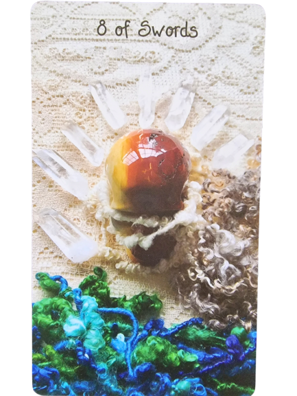 A tarot card from the Crystal Skull Tarot, titled 8 of Swords, features a central stone surrounded by eight clear crystals on a textured fabric background, with blue and green yarn at the bottom.