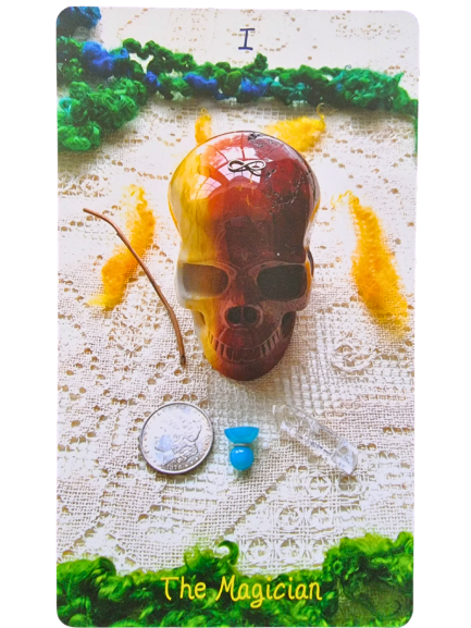 A Crystal Skull Tarot card titled The Magician displays a red and yellow skull, a coin, a small blue object, a clear crystal wand, and a stick arranged on lacy cloth with green and yellow decorations.