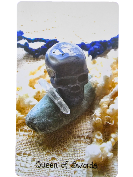 A tarot card labeled Queen of Swords from the Crystal Skull Tarot features a dark skull-shaped stone, clear quartz crystal, and an oval green stone, arranged on textured fabric with blue and yellow yarn in the background.