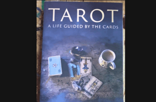A copy of Tarot: A Life Guided by the Cards by Maddy Elruna rests on a surface, its cover adorned with tarot cards, a key, and a cup of tea. "Book Review" is prominently displayed above the captivating scene.