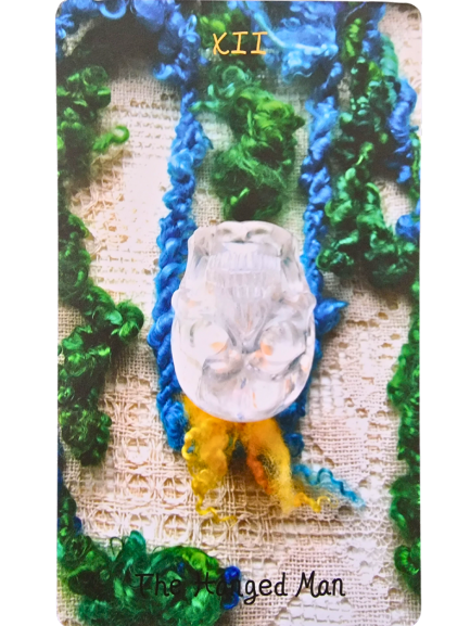 A Crystal Skull Tarot card titled The Hanged Man (XII) features a white, sculpted face upside down, with blue, green, and yellow yarn strands on a textured, lace-like background.