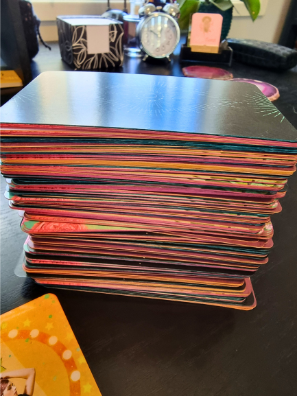 A large, colorful stack of blended decks with various designs on the edges sits on a dark tabletop. In the background are a clock, a plant, and other small objects, slightly out of focus.