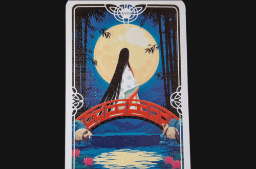 Tarot of the Divine’s The Moon card shows a figure in a kimono on a red bridge beneath a full moon, two white animals nearby, and water with lotus flowers below. Deck Review is displayed above.