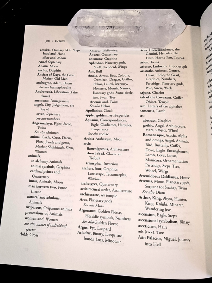 An open book, titled A Dictionary of Symbols, displays an index page listing words alphabetically in three columns. A large, clear quartz crystal rests on the top edge of the book. The text and crystal are clearly visible.