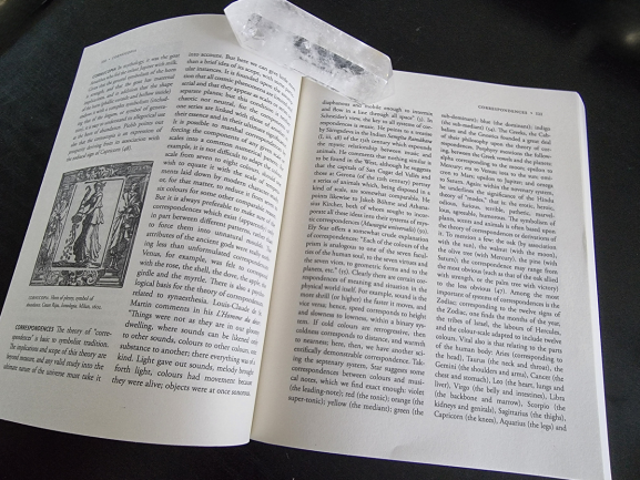 An open book, possibly A Dictionary of Symbols, lies on a dark surface held open by a clear quartz crystal. The left page features an illustration of two women with a column, and both pages are filled with dense text.