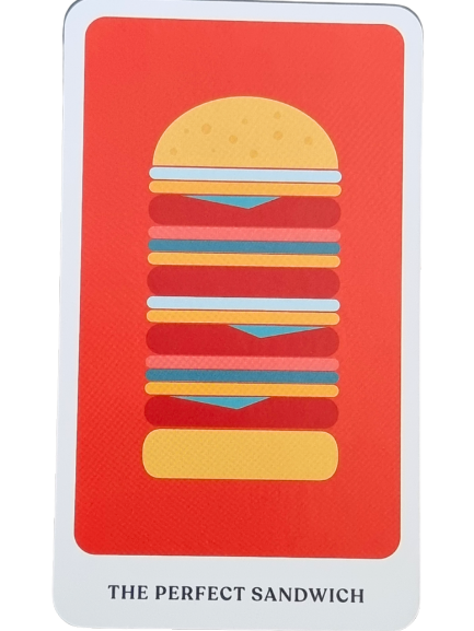 A playing card from The Story Oracle features a stylized illustration of a tall sandwich, stacked with bread, meat, cheese, and vegetables on a red background. Text at the bottom reads THE PERFECT SANDWICH.