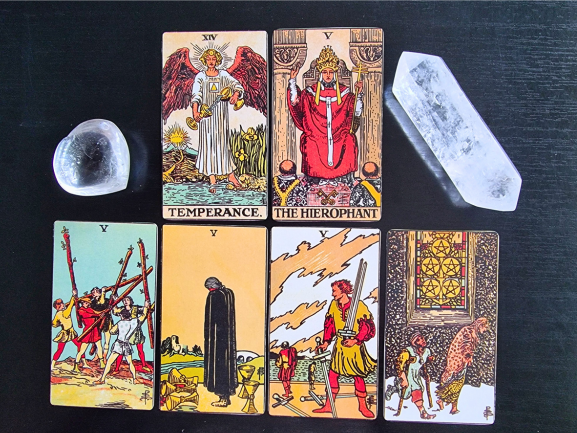 A tarot card spread for your personal year, featuring Temperance, The Hierophant, the Fives, and accented with a heart-shaped stone and clear quartz crystal on a black surface.