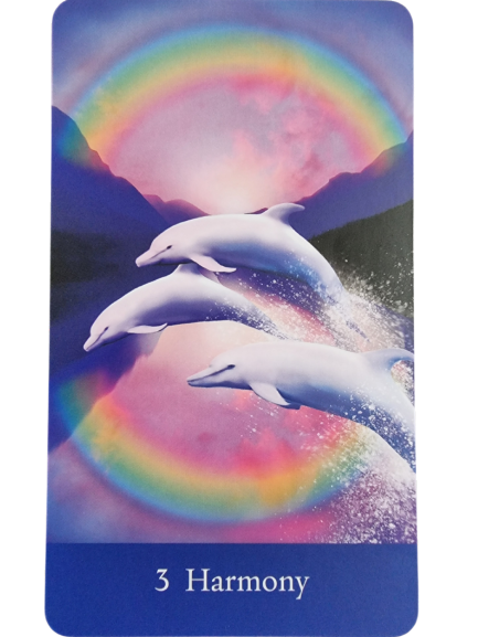 Three dolphins leap through a reflective body of water beneath a vibrant rainbow, framed by purple mountains and a pink sky. Inspired by The Sacred She Tarot, the word Harmony and the number 3 appear at the bottom.